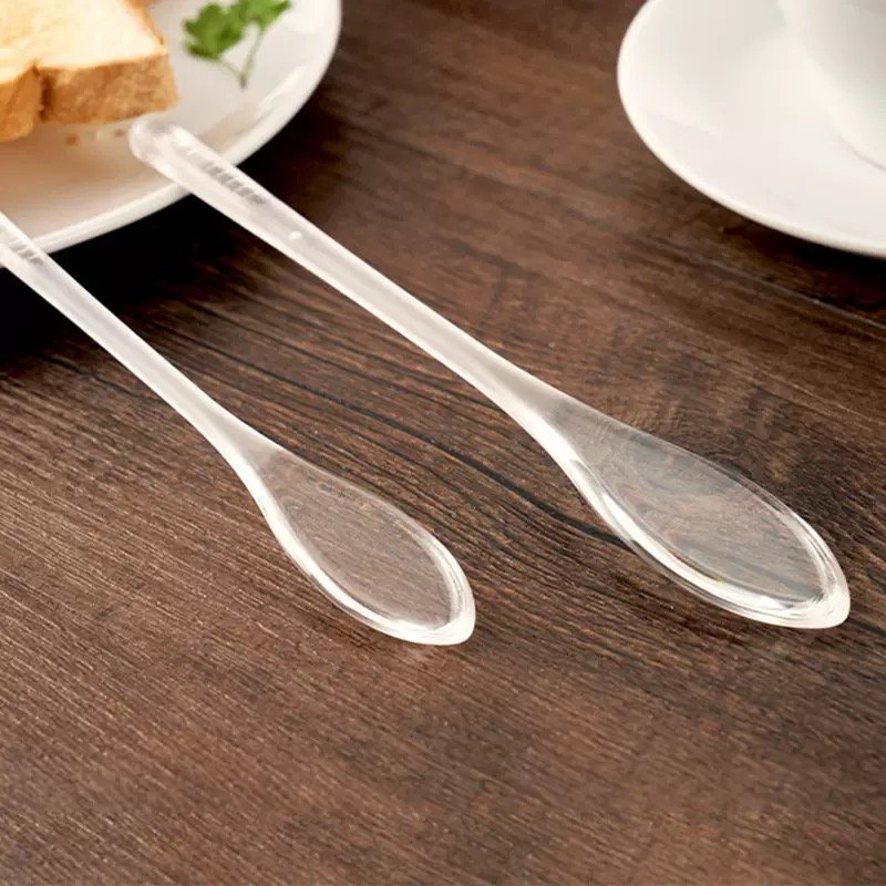 ZEST clear plastic honey dipper stick spoons with flat leaf-shaped heads on a wooden table beside breakfast toast